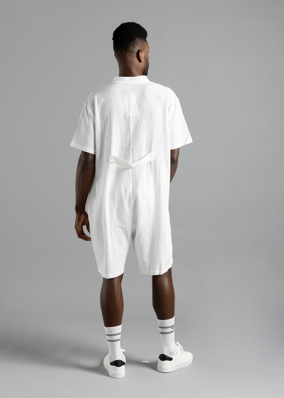 Rear view of the white MW Button Up Jumpsuit by MilkWhite on a model, showing the short sleeves and buttoned waist strap.