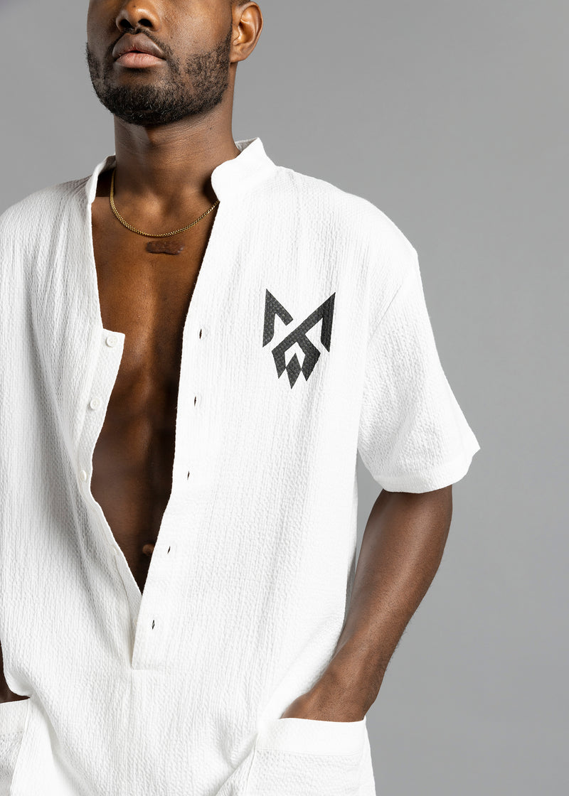 Close-up of the white MW Button Up Jumpsuit by MilkWhite, showing the black chest logo, buttons and textured surface.