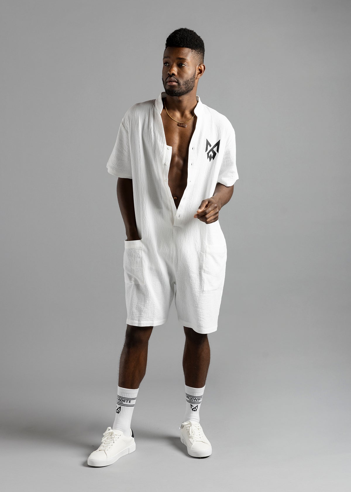 Front view of a model looking to the side in the white MW Button Up Jumpsuit by MilkWhite, showing its black chest logo.