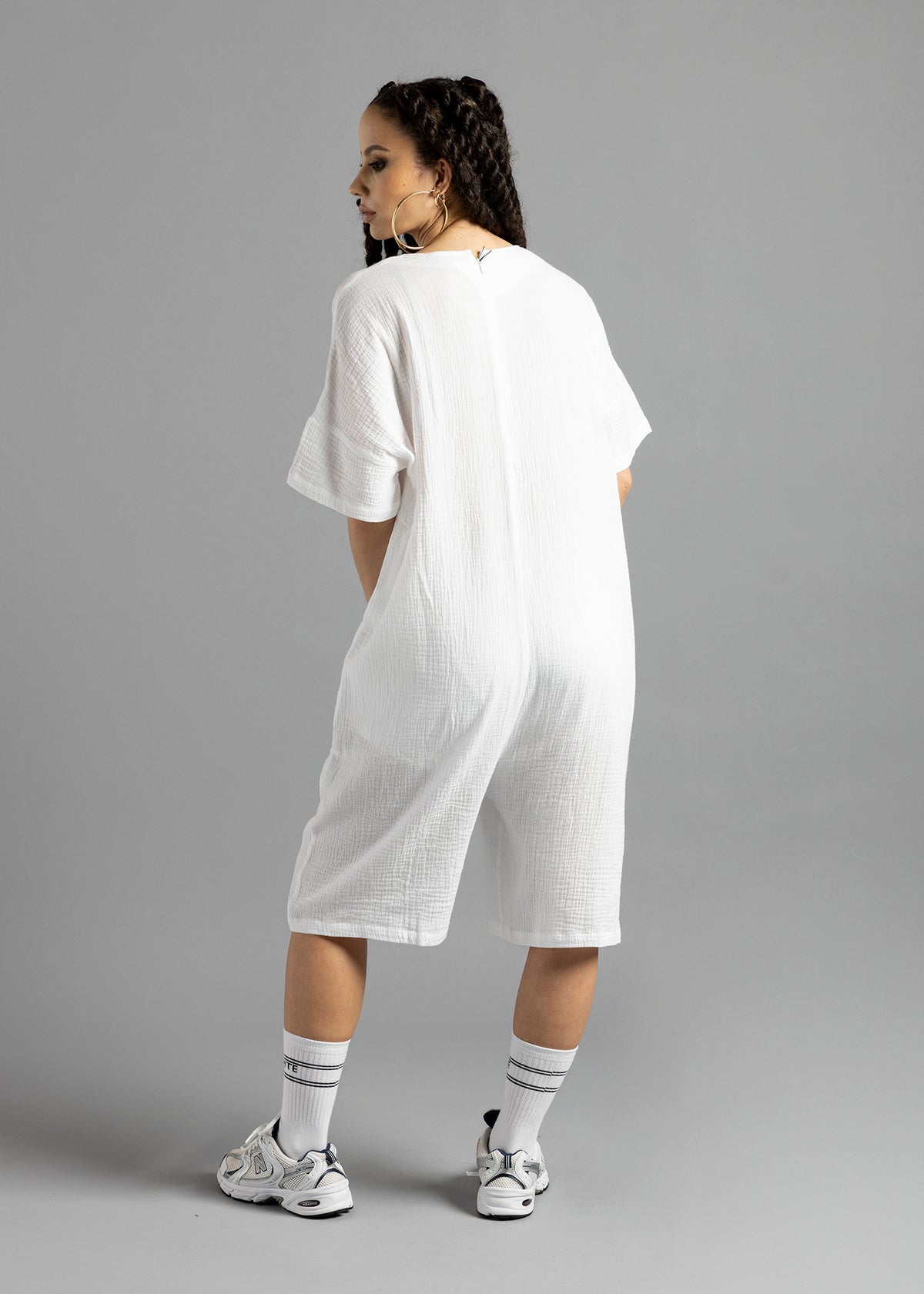 Full-length rear view of a model wearing the white MW V-Cut Jumpsuit by MilkWhite, showing its plain back, short sleeves and knee-length legs.