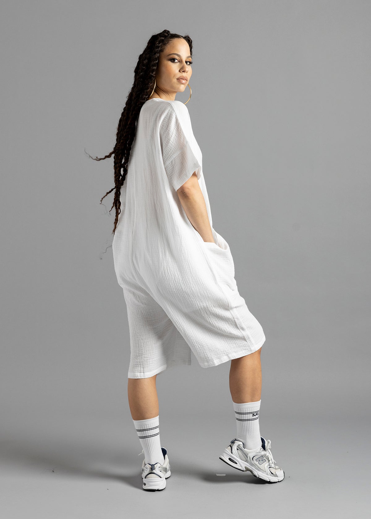 Three-quarter rear view of a model wearing the white MW V-Cut Jumpsuit by MilkWhite, with one hand in a pocket and head turned toward the camera.