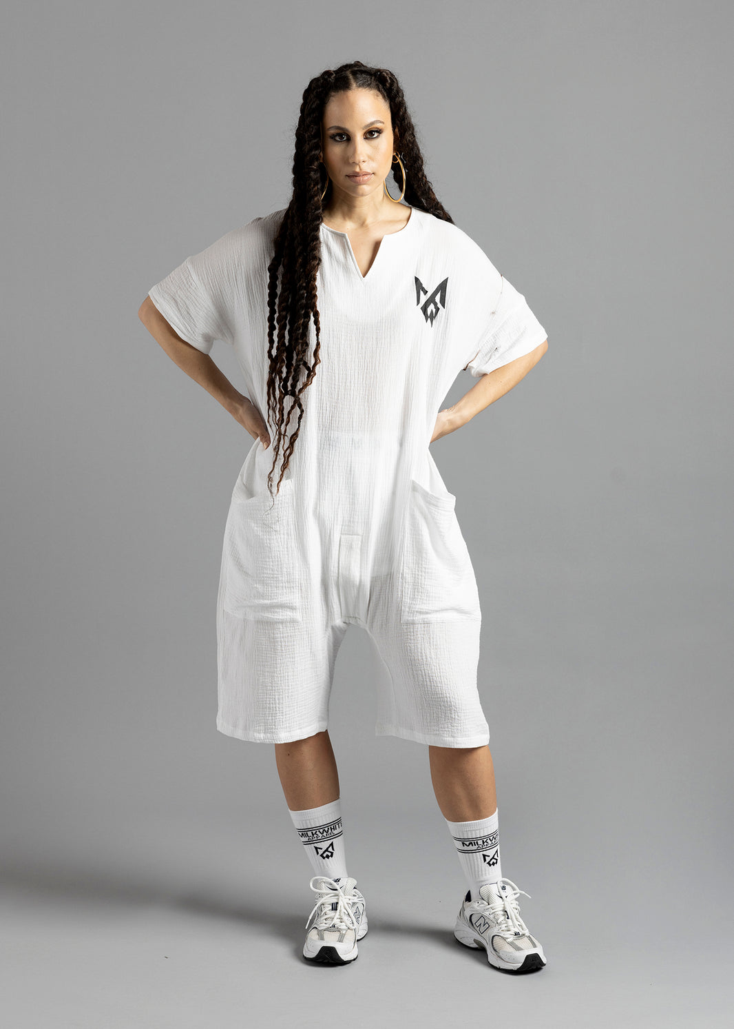 Front view of a model wearing the white MW V-Cut Jumpsuit by MilkWhite with hands on hips, showing the V-shaped neckline and black chest logo.