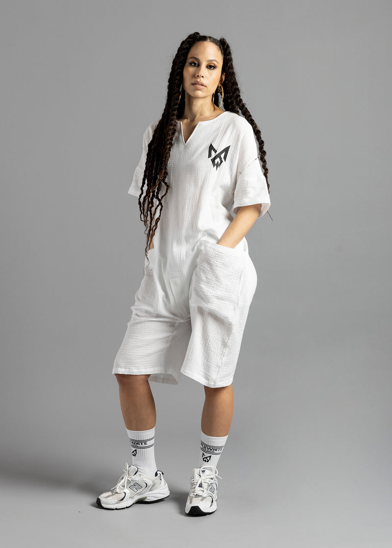Three-quarter front view of a model wearing the white MW V-Cut Jumpsuit by MilkWhite, showing the chest logo, front pocket and knee-length legs.