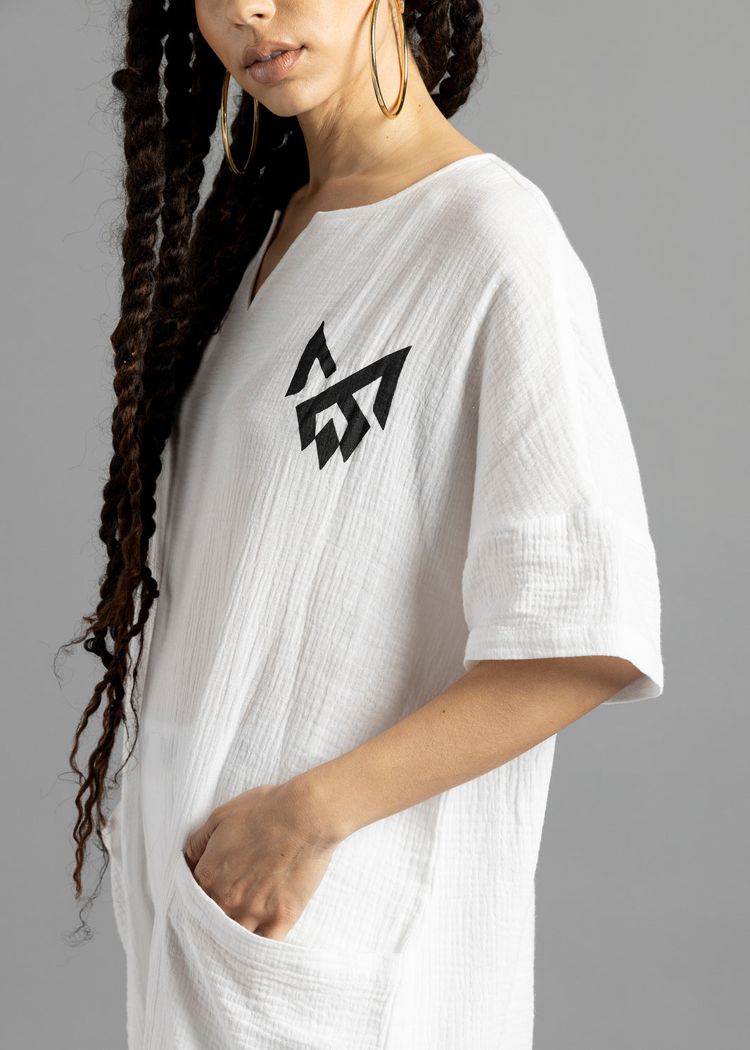 Close-up of the white MW V-Cut Jumpsuit by MilkWhite, showing the black chest logo, textured fabric and a hand in the front pocket.