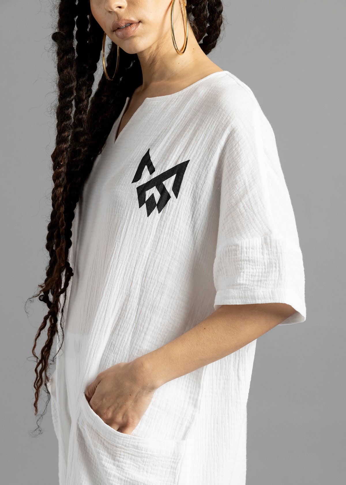 Close-up of the white MW V-Cut Jumpsuit by MilkWhite, showing the black chest logo, textured fabric and a hand in the front pocket.