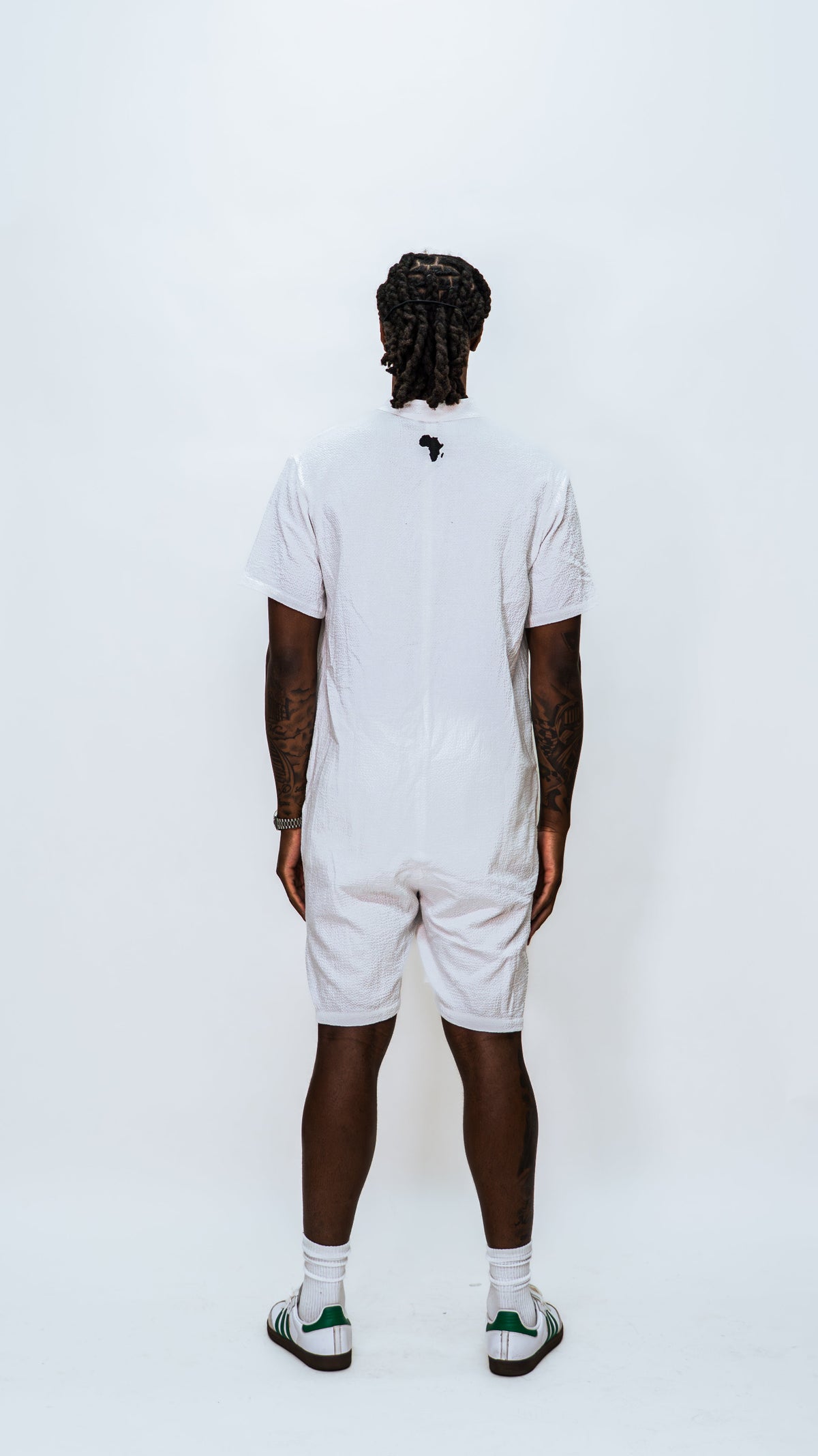 Full rear view of a model wearing a Milkwhite white jumpsuit with a black silhouette logo of africa near top of the neck.