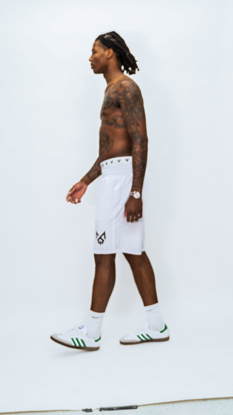 Model walking in white MilkWhite Long Swim Trunks, shown in side profile with the black logo visible near the hem.