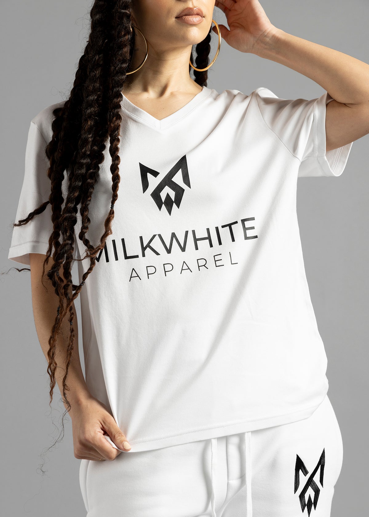 Front close-up of the white MilkWhite Premium T-Shirt on a model holding the hem, showing the V-neck and dark chest logo and lettering.
