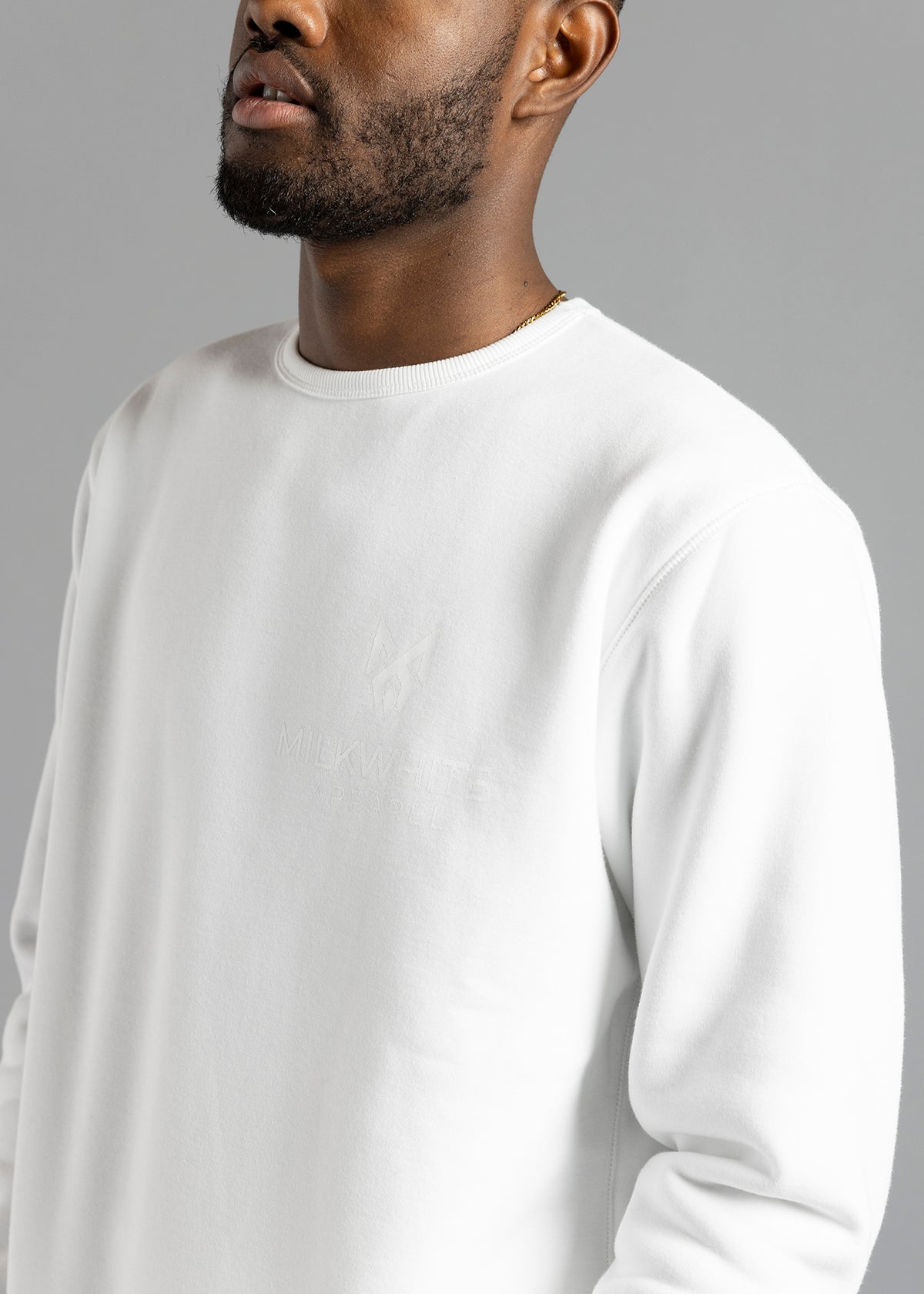 Chest close-up of the white Milk Essential Sweatshirt by MilkWhite, showing the white-on-white logo and lettering beneath the ribbed crew neckline.