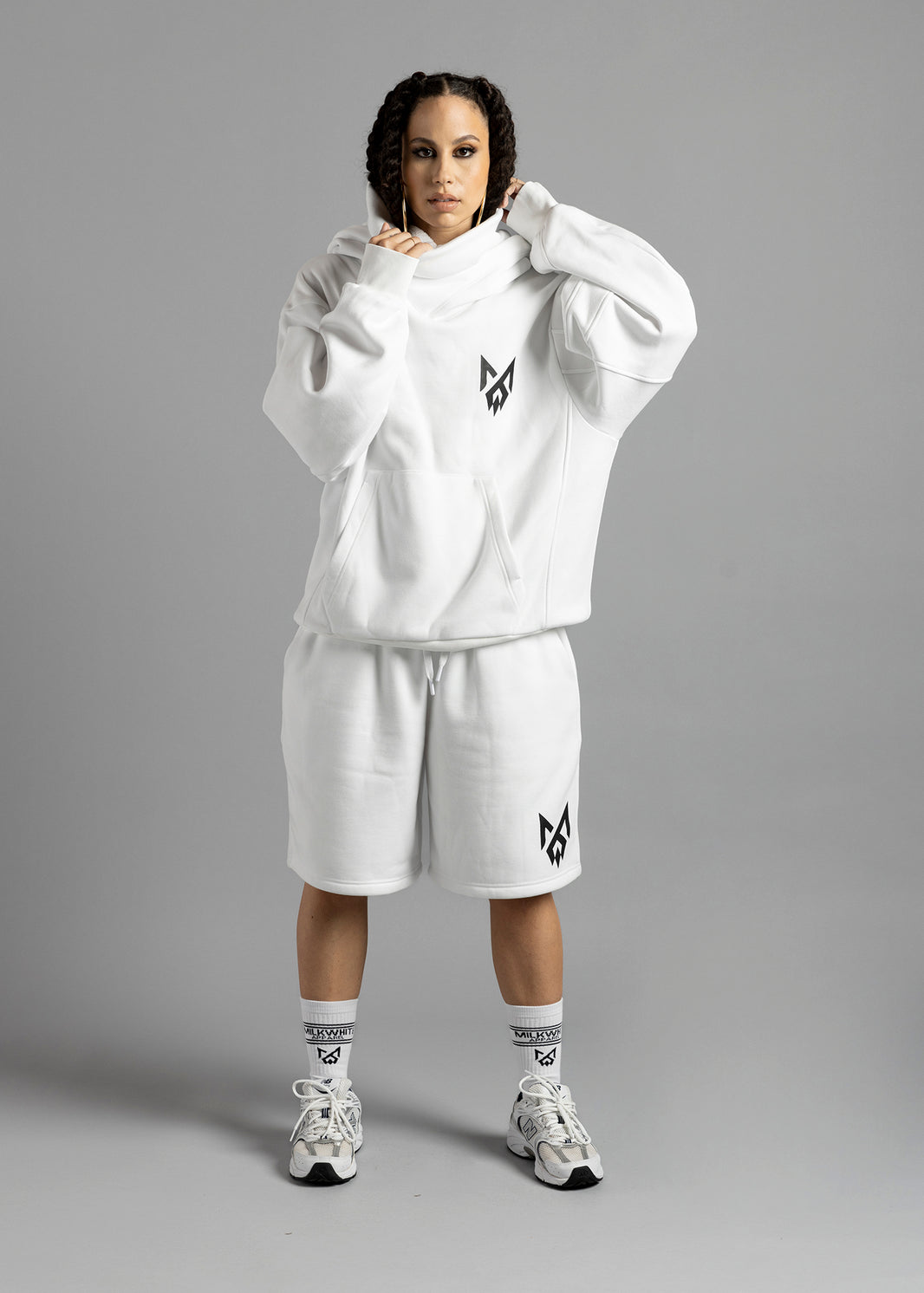 Full-length front view of a model wearing the white Rosebud Hoodie by MilkWhite, holding the draped neckline above the black chest logo.